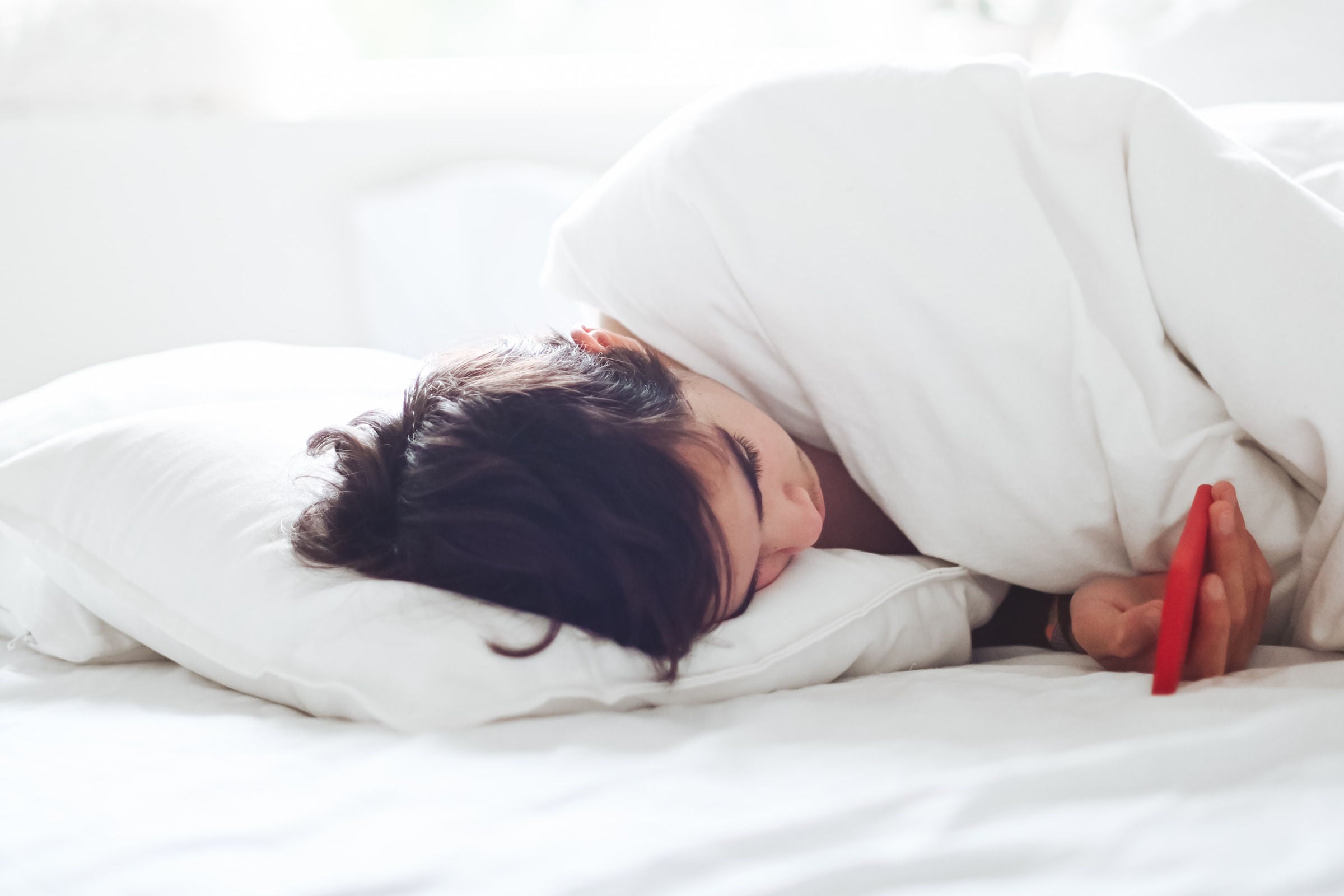 A woman lying in bed looking at her phone while tucked in a white blanket.