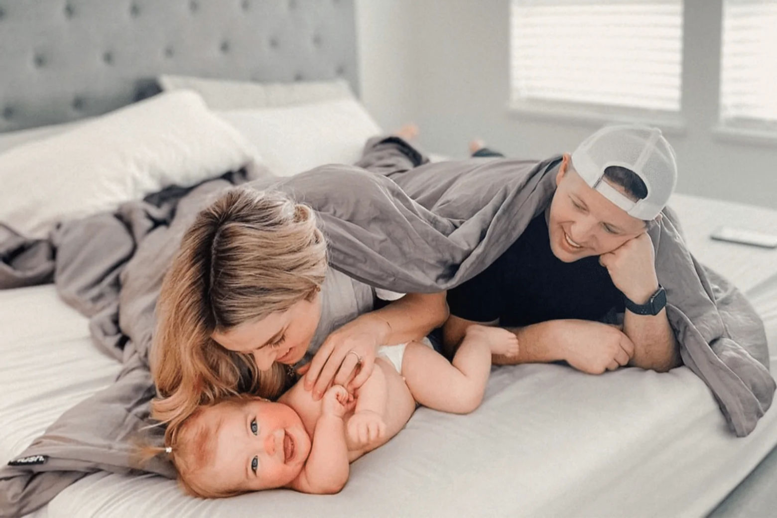 A happy family playiong with their baby while under a Hush Iced Cooling Weighted Blanket in bed.