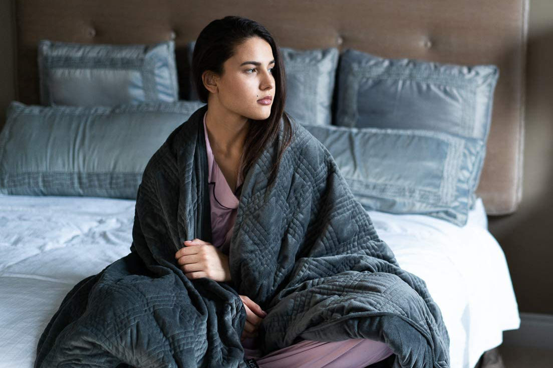 A young woman sits comfortably on bed while covered in a grey Hush classic weighted blanket.