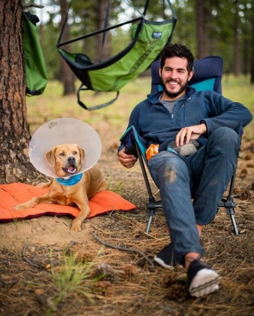 A man wearing Vessi Storm Black Cityscapes while sitting in a camp chair next to his dog out in the wilderness