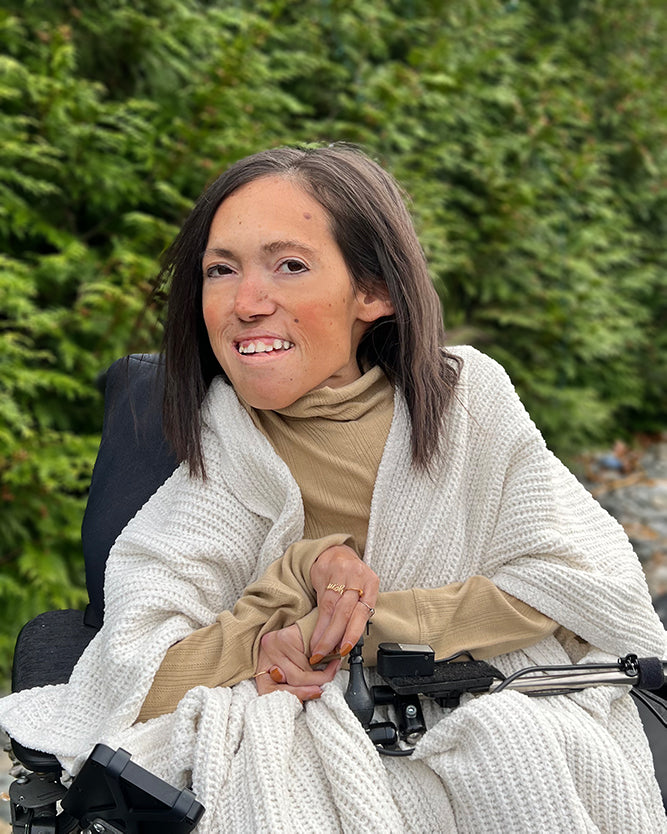 Alyssa Silva sits outside in her powerchair against a backdrop of foliage, wearing a knit shawl and a beige turtle kneck.