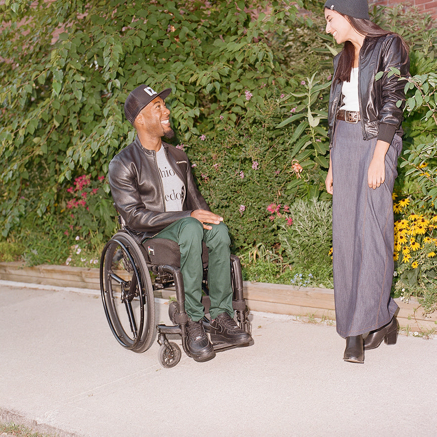 A handsome man with a big smile wearing green chinos and a black IZ Adaptive leather jacket sits in a manual wheelchair looking up at a beautilful woman who is standing, wearing an IZ Adaptive toque, leather jacket, and an IZ standing denim skirt. She is smiling back at him as they stand in front of green foliage.