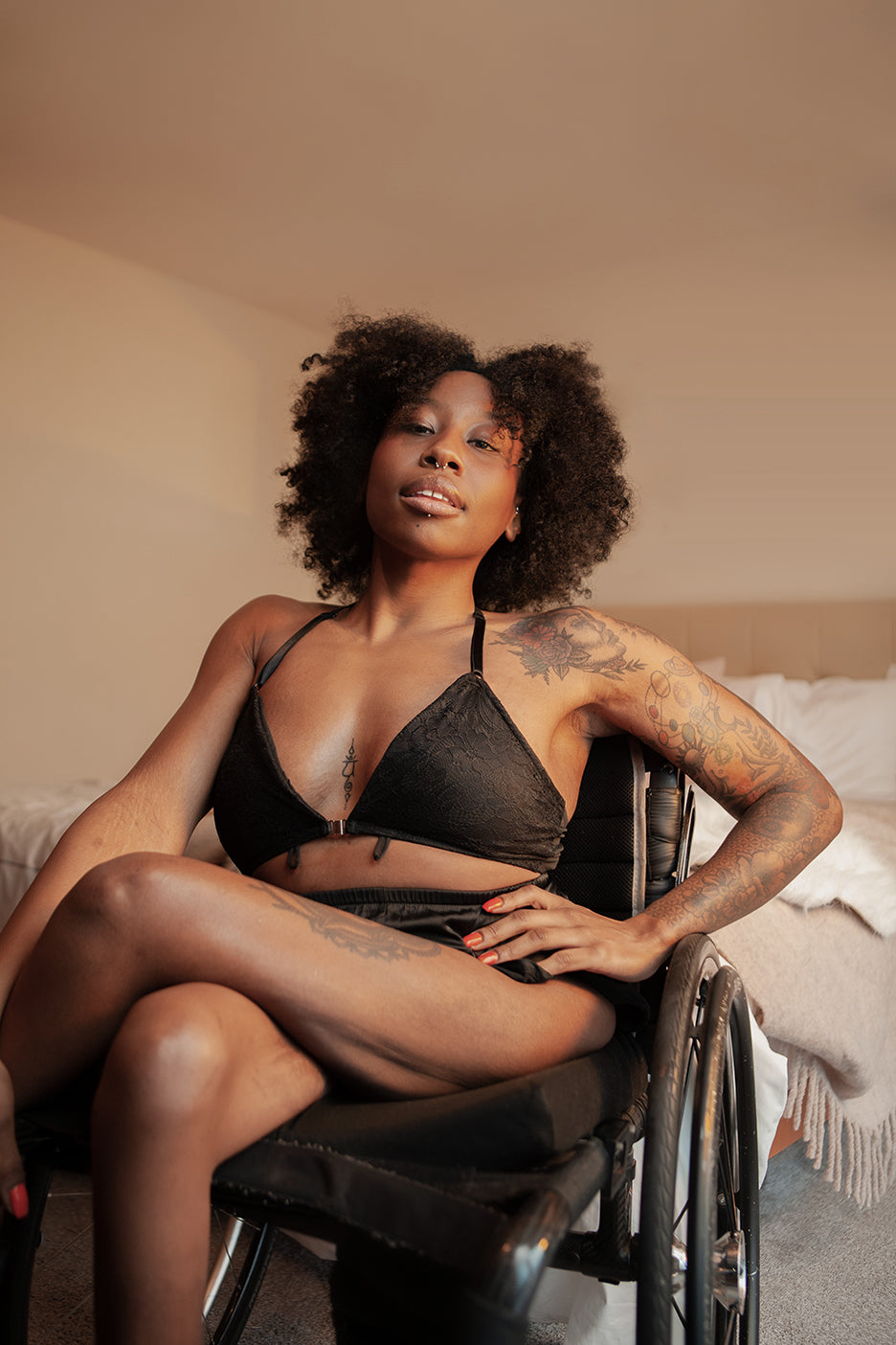 A Liberare model sits in her manual wheelchair wearing very sexy Liberare underwear and bra, with her hand on her hip and her legs crossed. She's in a bedroom with a bed behind her and beige walls.