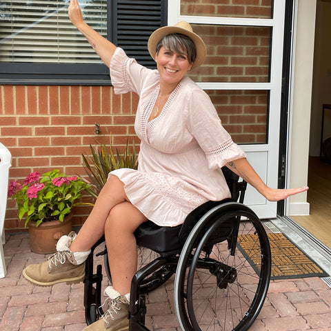 Sandie Roberts sits in her black wheelchair with her arms outstretched and her legs crossed. She has a big smile. She is wearing a light pink flowy summer dress, a straw brimmed hat, and beige suede lace-up boots. She is outside on a brick patio, in front of a brick wall with some flowers behind her.