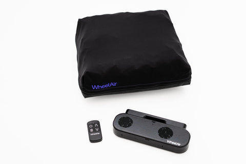 A black Wheelchair cushion with a small remote control and the WheelAir system, a double bladed fan encased in a black plastic housing in this case, lying n a flat white background.