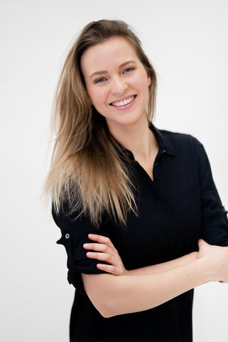 Corien Staels stands in front of a grey background wearing a button up black blouse rolled up to her elbows. Her arms are crossed and she has a big smile.