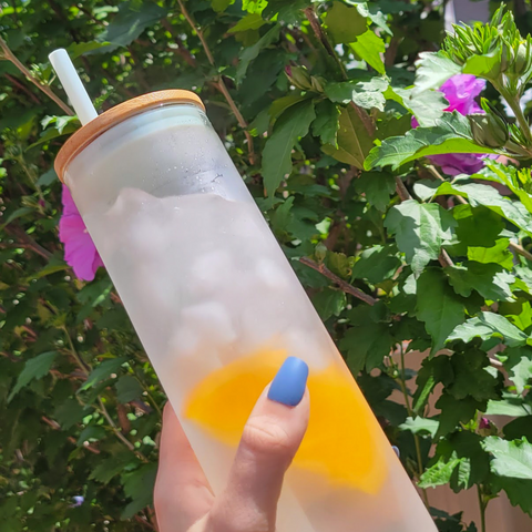 Hand holding a glass water bottle with bamboo lid and straw with ice and orange infused water