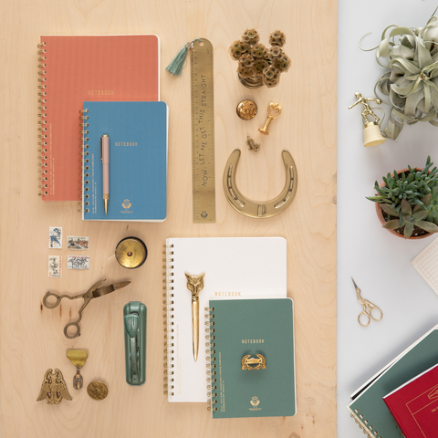 Flatlay of large pink notebook, small blue notebook, large white notebook, small green notebook, scissors, rulers, and more office accessories on a wooden desk with gold desk accessories and trinkets around the spiral notebooks