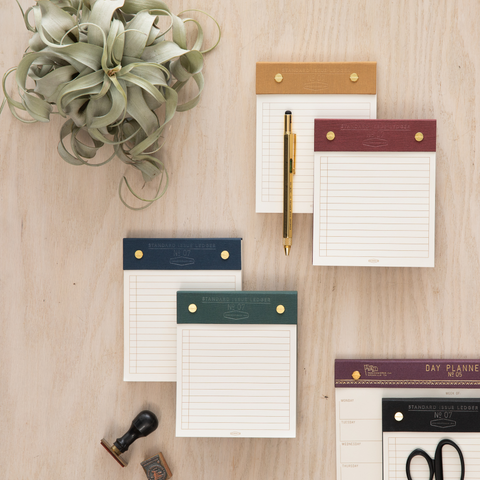 Four Standard Issue Post Bound Ledger Note Pads with Pen sitting on top on wooden desk with plant to the side