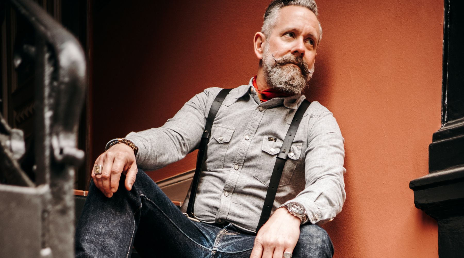 Man with beard Leaning against rust colored wall wearing Black Leather Wiseguy Original Suspenders and a grey canvas western style shirt and jeans