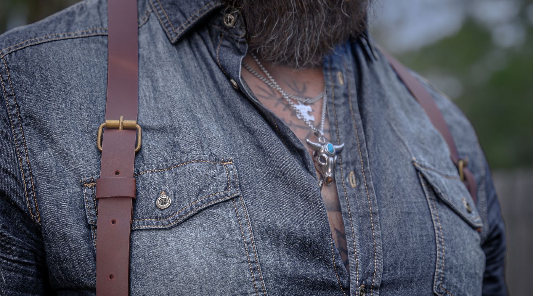 Close up view of man wearing red leather Wiseguy Original Suspenders with brass buckles. He is wearing a denim western style shirt and silver necklaces with a turquoise stone.