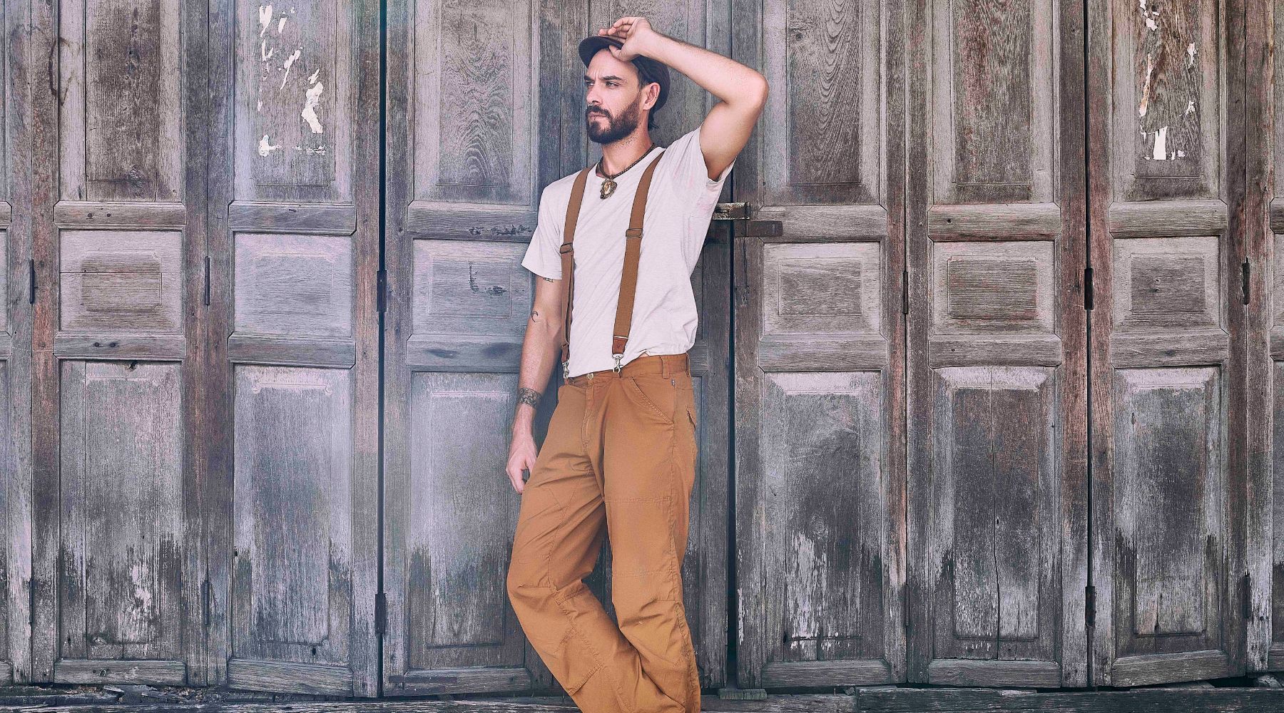 Man with flat cap and a white t-shirt and mustard colored pants is wearing Duck Cotton Wiseguy Original Suspenders in the color Mocca. He is standing in front of a big old wooden discolored door.