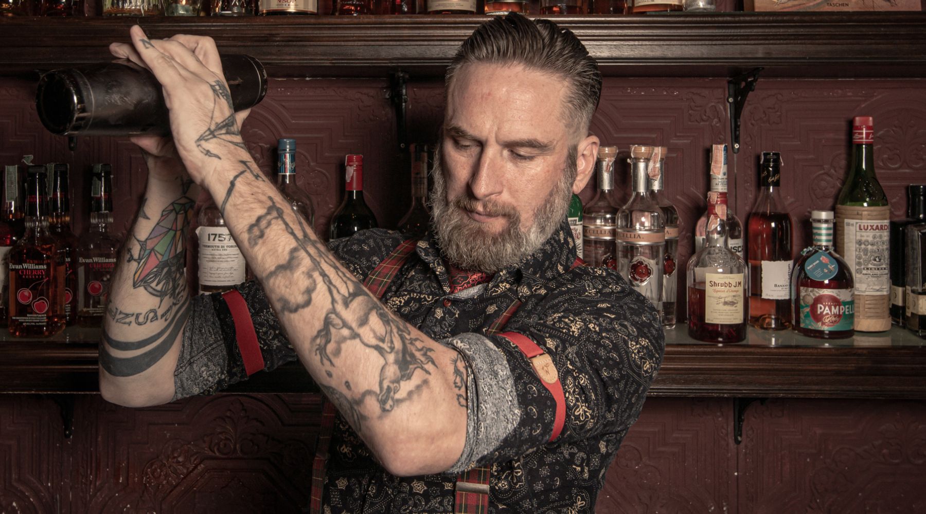 Man standing behind bar with a cocktail shaker in his hands, he is wearing a patterned shirt with red Wiseguy Original Sleeve Garters and Suspenders. He has a beard and Mustache.