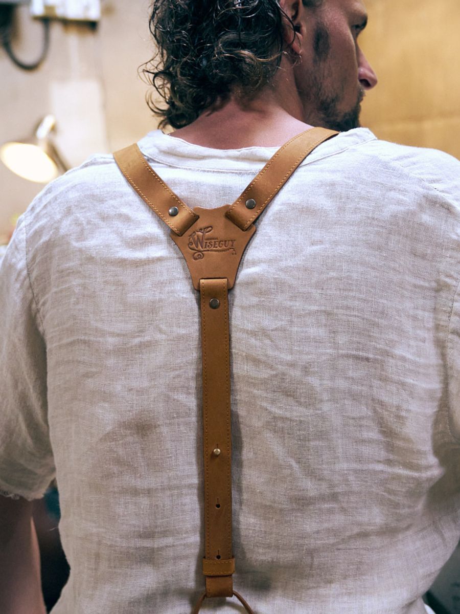 Mann mit Locken auf dem Rücken, trägt ein weißes Leinenhemd und kamelfarbene Hosenträger von Wiseguy Original mit Nähten. Das Logo von Wiseguy Original befindet sich in der Mitte des Bildes.