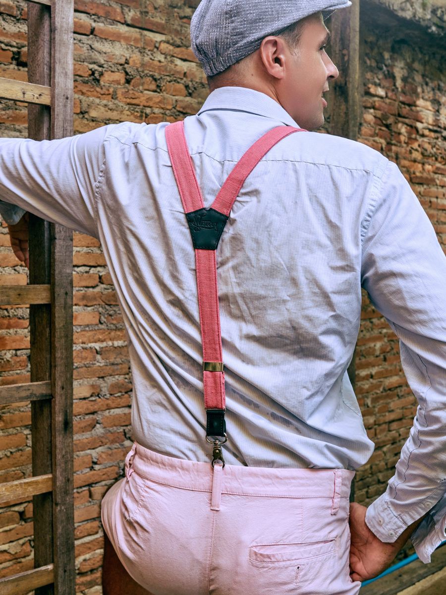 Man from the back wearing a flat cap, a light blue shirt and pink linen suspenders with black leather. The man is wearing pink shorts and is leaning with his left arm on a brick wall.