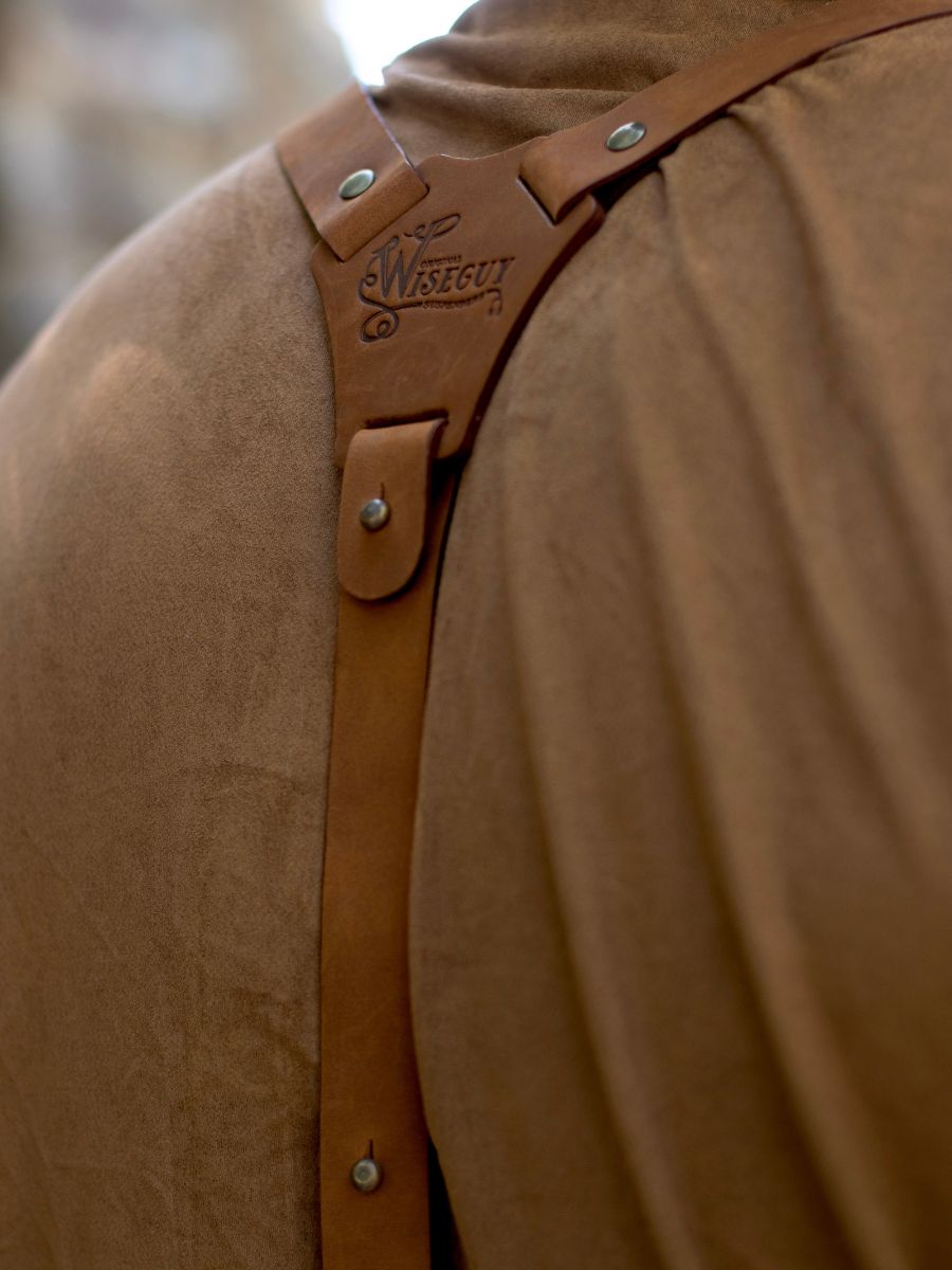 Close up shot of someone's back wearing camel colored leather Wiseguy Original Suspenders, they have a brown t-shirt or shirt on. The Wiseguy Original logo is the part of the picture that is in focus.