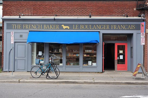 Image of the front facade of The French Baker storefront. The building is painted medium grey, with a cyan blue canopy extending into the sidewalk from over the windows. The double entry doors are painted bright red.