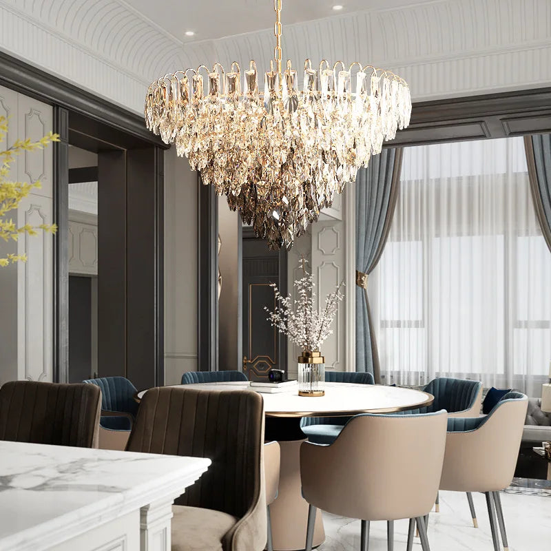 A modern dining room features a round table, beige and blue chairs, and the Morsale Langford Luxury Modern Chandelier overhead. Tall windows with blue curtains fill the space with light, highlighting the stylish crystal fixture above.