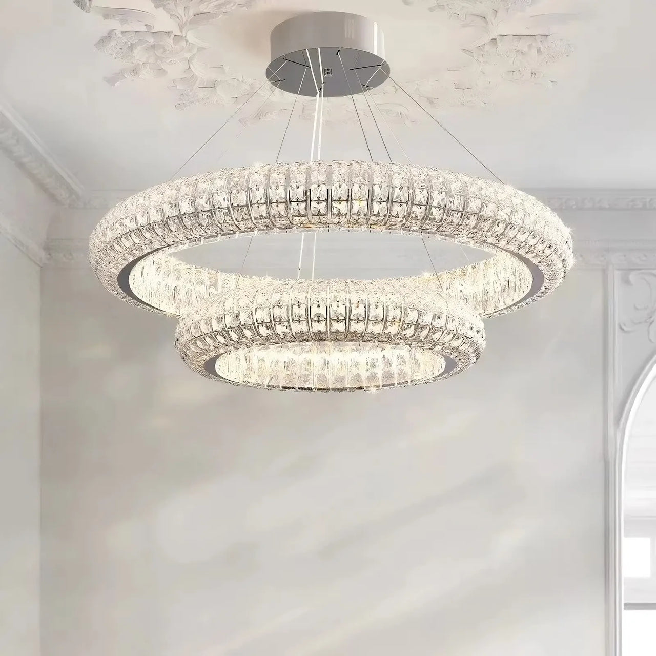 The Bacci Crystal Modern Chandelier by Morsale, a luxurious two-tiered fixture with a polished chrome finish and sparkling crystals, hangs in an elegantly decorated room with intricate molding and a partial view of an arched window on the right.