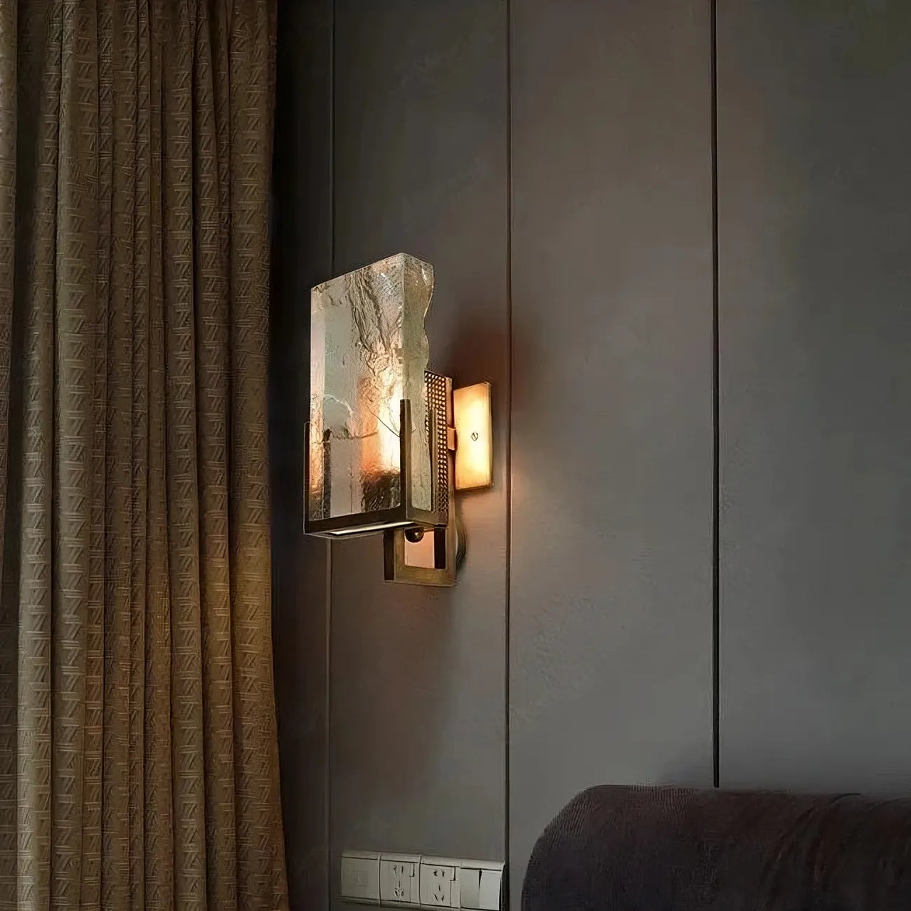 The Palermo Crystal Wall Sconce by Morsale.com features a modern rectangular crystal shade, casting a warm glow on grey walls near brown curtains and above a dark sofa, with outlets below—ideal for both modern and traditional spaces.