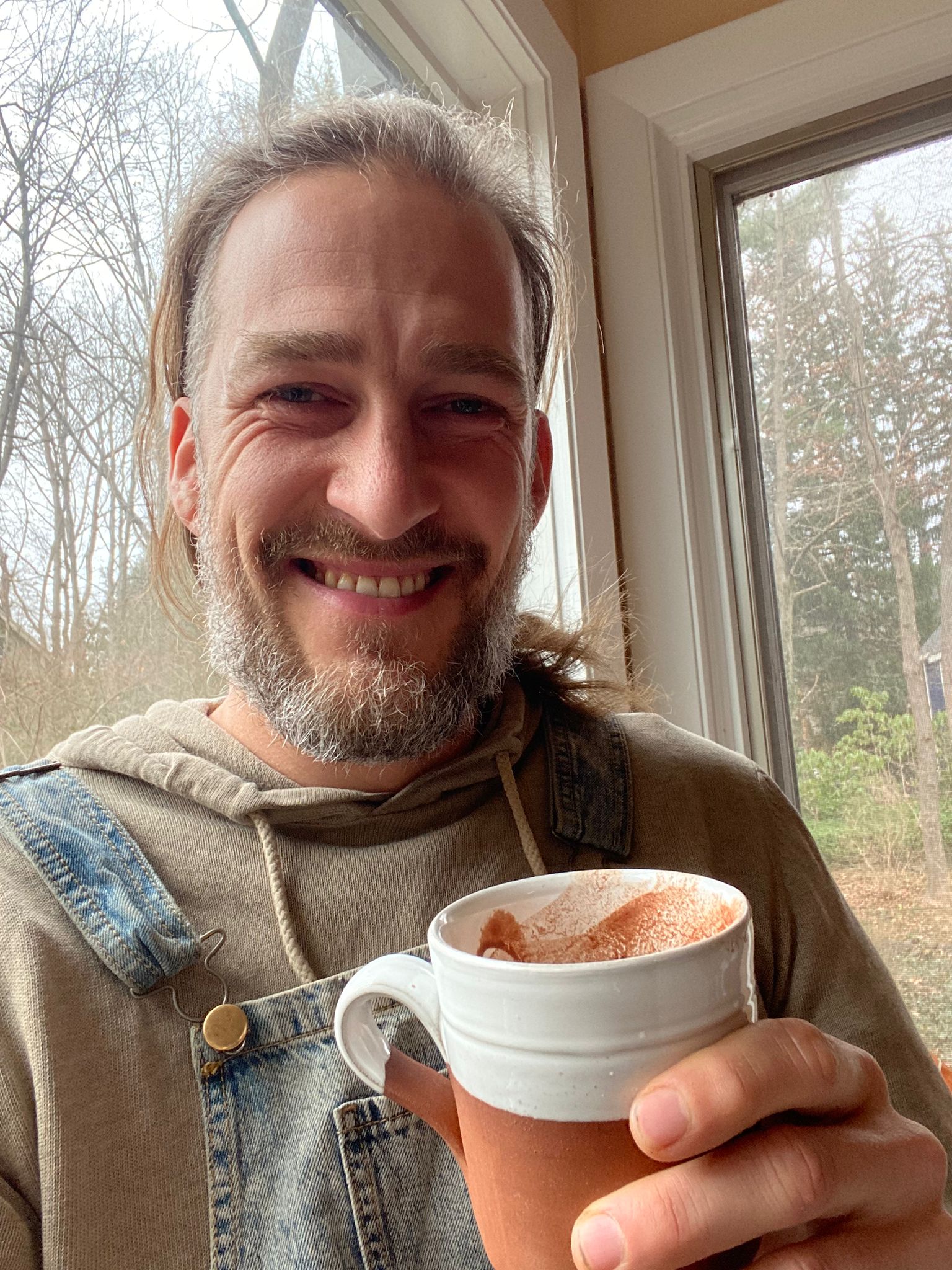 Man smiling holding a ceramic mug by a window with a forest view.