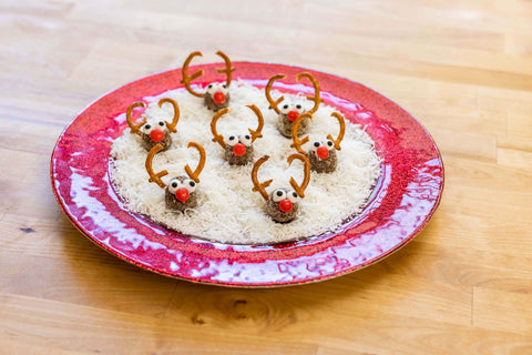 A plate of Frooze Balls dressed as Rudolph with broken pretzels, googly eyes, and red noses. The plate is covered in coconut flakes to resemble a layer of snow. 