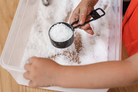 Kid holding messy play materials including a plastic bin and measuring cup.