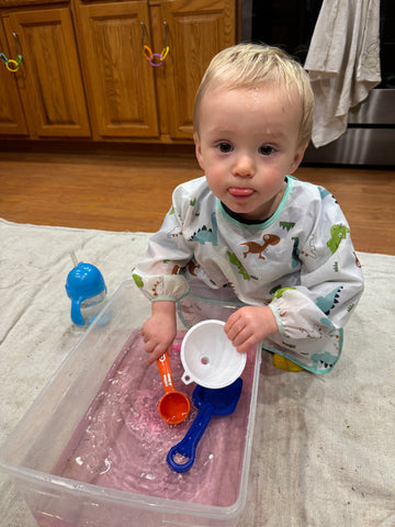 Baby plays with pink water in a plastic tub with toys for Valentine's Day.