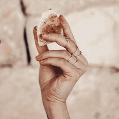 White female hand holding Citrine crystal and wearing gold rings against a stone wall background