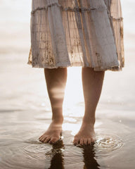 Woman's legs standing in shallow sea water wearing a floaty dress with sun shining on the water's reflection.