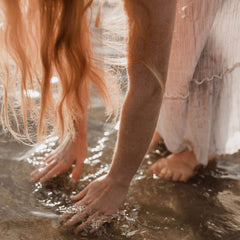 Woman in floaty dress standing in shallow sea water bending forwards with hands touching the sea and hair falling towards the water.