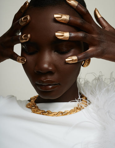 Black model with gilded fingertips in a white top with gold jewelry.