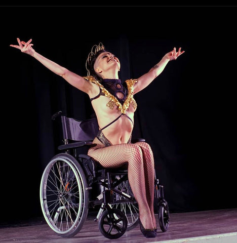 little peaches is in a wheel chair dressed in gold with a flamboyant headpiece, she has her arms spread wide and is smiling to the audience. 
