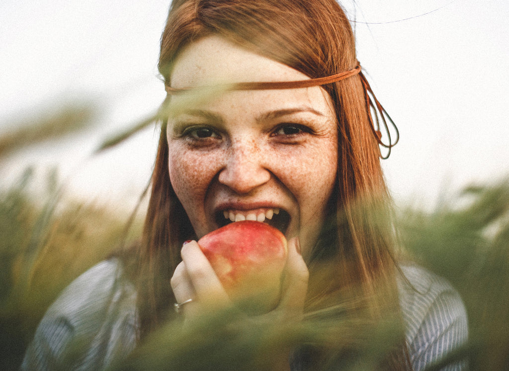 Happy long-haired young woman outdoors biting a red apple which could make her mouth tingle if she has birch pollen allergy