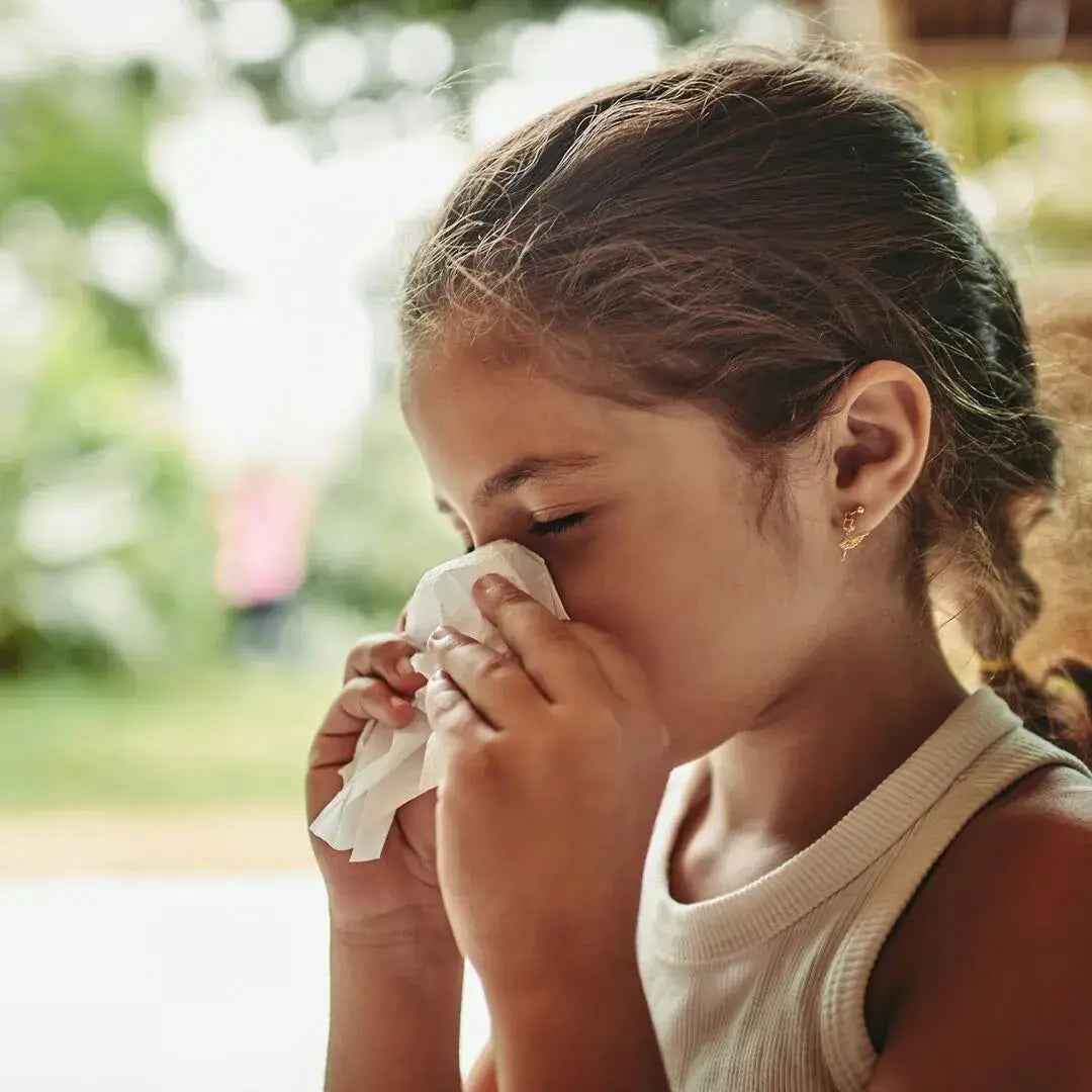 Child sneezing into a paper tissue and blowing her nose – she’s in a summer dress standing by a window