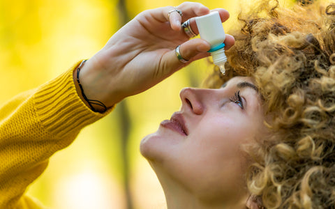 Stylish curly-haired woman in a yellow sweater squeezing drops into her eyes made itchy and watery by grass pollen allergy