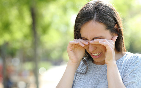 Woman rubbing her eyes outside under the trees – itchy, red or watery eyes are a common symptom of seasonal allergies