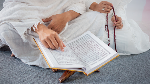 Two people sitting cross-legged, dressed in lightweight cotton clothing.  There is an open Quran on a Quran stand, with one person reading along, using the index finger to follow the text, and the other person is holding tasbih beads.
