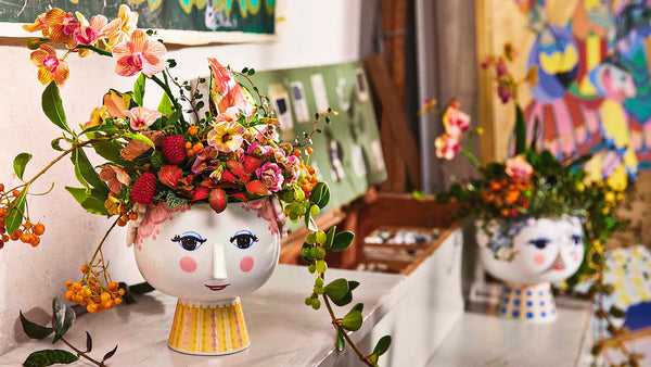 Eva flowerpot is made of glossy glazed ceramic with coloured decal and is inspired by Wiinblad's original EVA vases, which had wide heads with floral decorations and a cone-shaped body. The delicate face is from Wiinblad's original line and the dress pattern is a reinterpretation of Bjørn Wiinblad's illustrations.