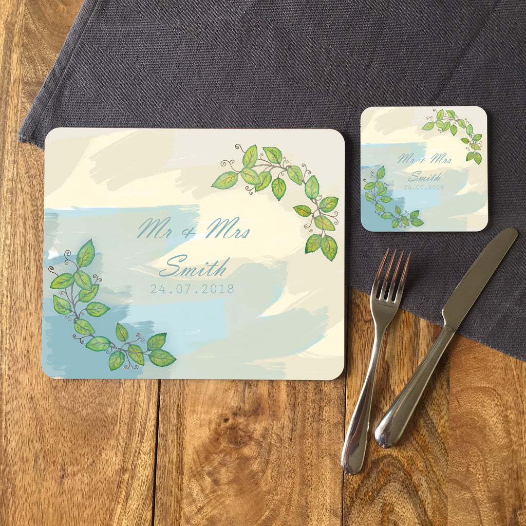 A personalised placemat and coaster on a table next to a knife and fork. The coaster and placemat feature a watercolour design with a leaf pattern