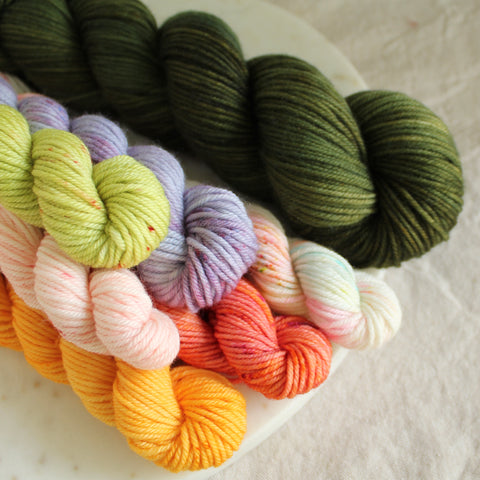 A skein of Deep forest green yarn sits on a marble plate with a pile of six brightly colored mini skeins