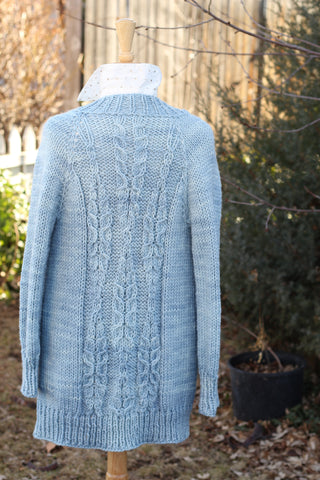 a long, hand knit cardigan with cables in the shape of fern leaves hangs on a mannequin near a spruce tree.