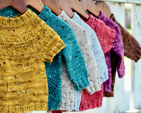 a row of gemstone colored sweaters hang on a fence. Gold, teal, sky blue, hibiscus pink, magenta, and chocolate