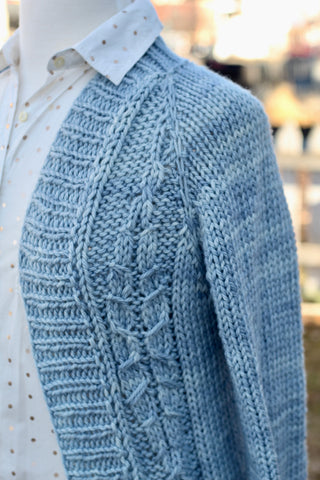 the front right side of a soft blue hand knit sweater is hanging on a mannequin over a white button down shirt with gold dots