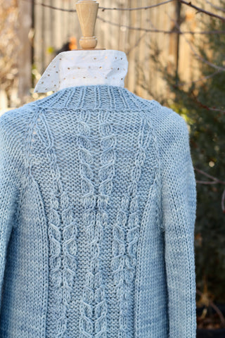 a sky blue hand knit cardigan with fern shaped cables in three vertical rows hangs on a mannequin