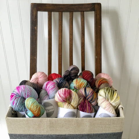 A linen basket of colorful, hand dyed yarn skeins sits on a wooden chair.