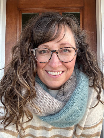 a smiling woman with curly brown hair and brown glasses is wearing a cowl that is soft tan and light teal.
