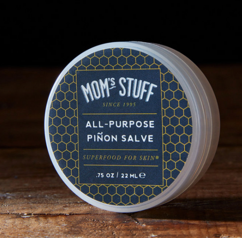 a round cansiter of salve sits on a wooden table. The label is navy blue with honeycomb and white lettering that says Mom's Stuff