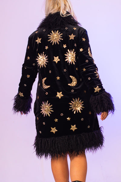 Woman modeling a black knee lenght boho style coat which features gold sequin stars and moons on the back.
