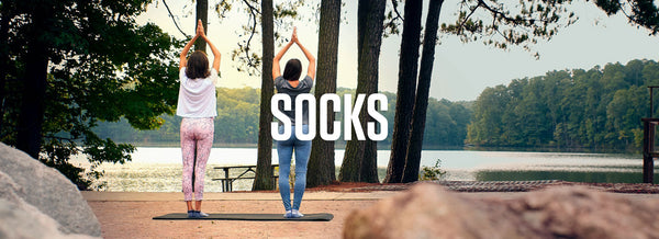 Sof Sole Socks Banner with two people on yoga mats in a park in front of a lake with trees in the distance doing a sun salutation.